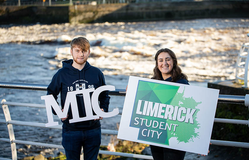 Two students holding a Limerick Student City sign in front of the River Shannon.