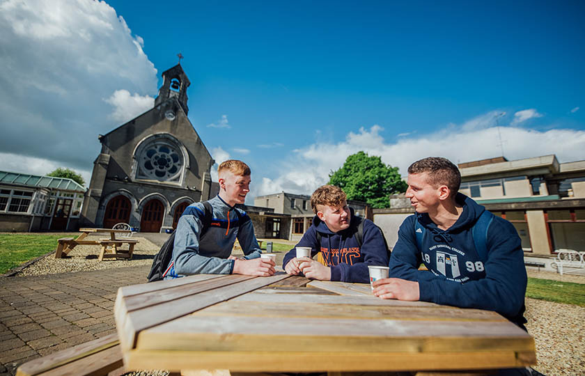 Three students sitting at a table in the quad wearing MIC branded hoodies and sportswear.