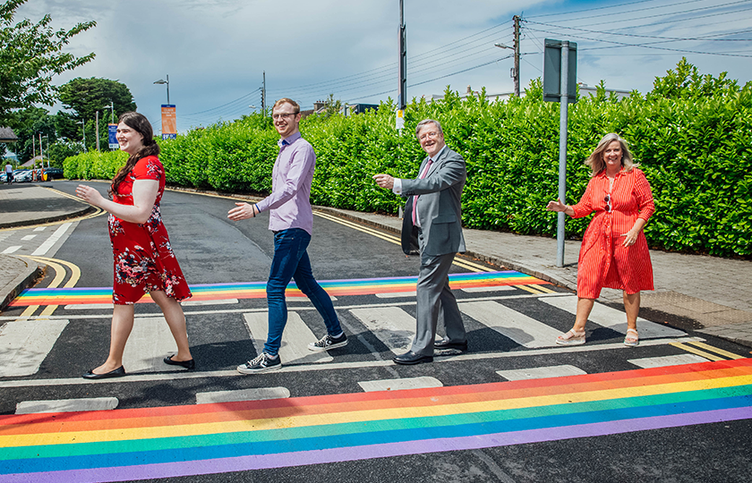 Four people walk single file across a pedestrian crossing with rainbow trim