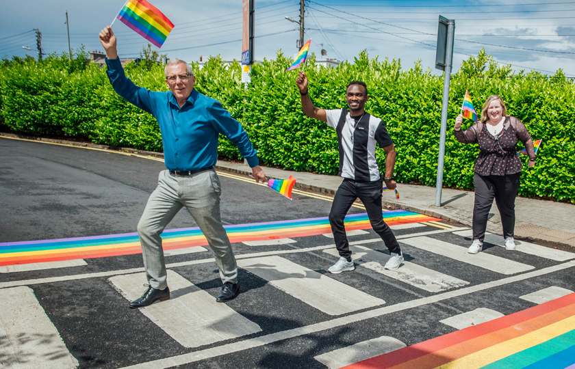Three people carrying small rainbow flags walk single file across a pedestrian crossing with rainbow trim