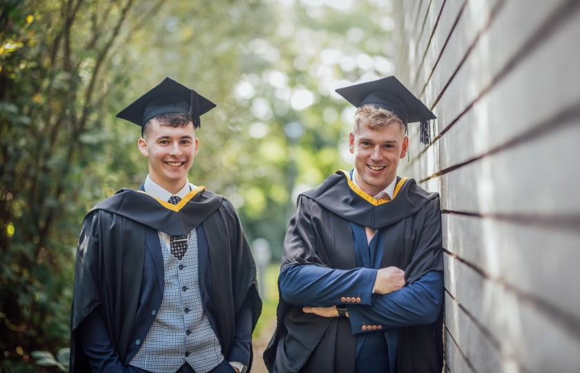 Two male graduates in their graduation gowns and hats outside MIC building.