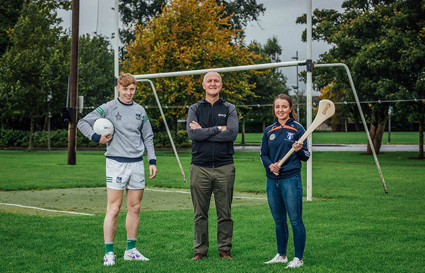 Pádraig de Brún holding a football, Ciarán Barr and Caoimhe Costelloe, holding a hurley in front of goals on sports field.  