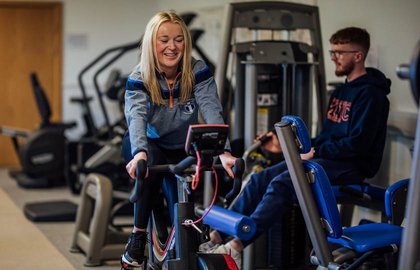 A female student with blonde hair is pictured on an exercise bike in a gym. There is a male student in the background.