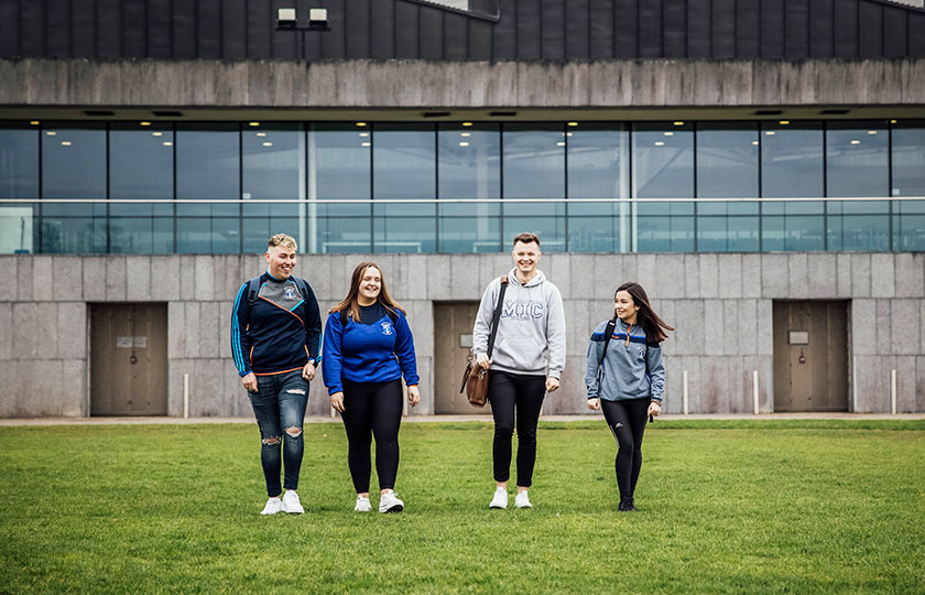 Four students walking across the playing field in front of MIC sports centre.