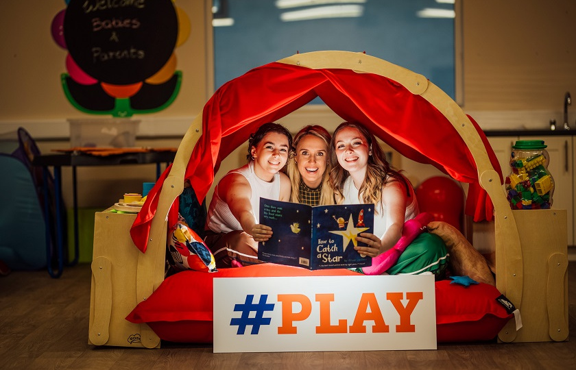 Three female people excitedly reading an illuminated book under cover of a red tent. A sign that reads "# play" sits in front of them.