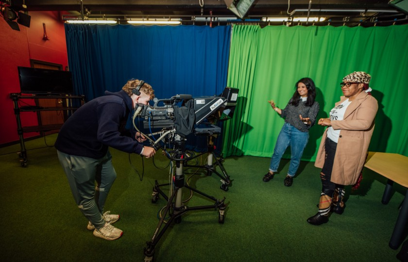 Male student behind a camera and two female students in front of camera in TV studio. There's blue and green screens set up behind them.