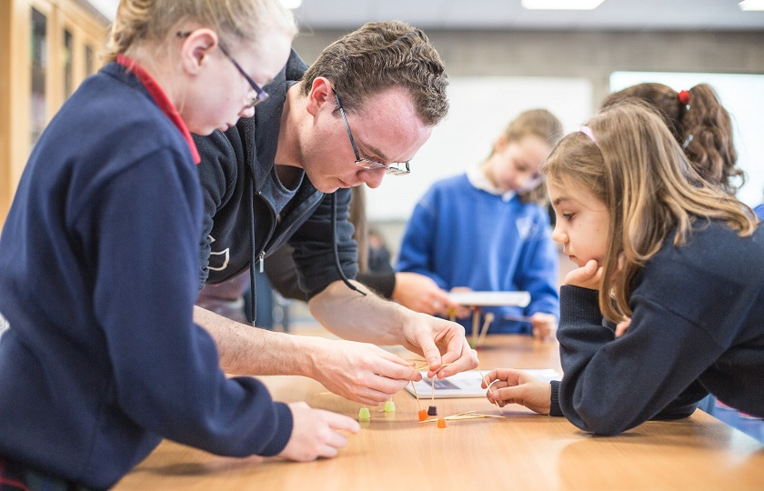 Male teacher helps two students, one male and one female, with a task at a desk