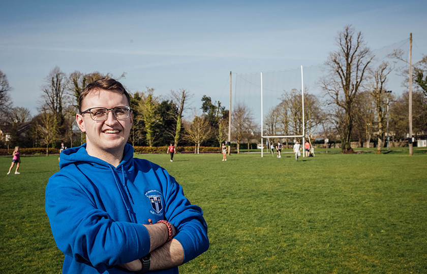 A student in an MIC hoodie standing on a grassy sports pitch.