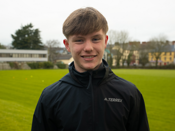 A male student is standing near a playing field. He is smiling at the camera and wearing black jacket.