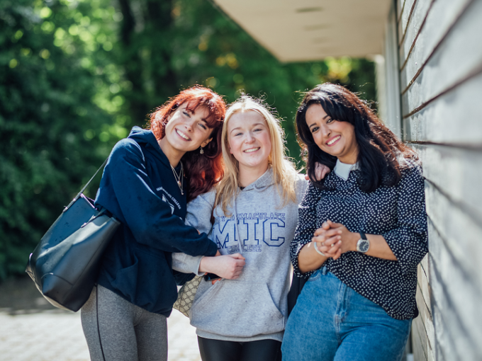 Three female students sanding together with their arms linked and looking at the camera. Two students are wearing MIC branded hoodies and the other student is wearing a polka dot top.